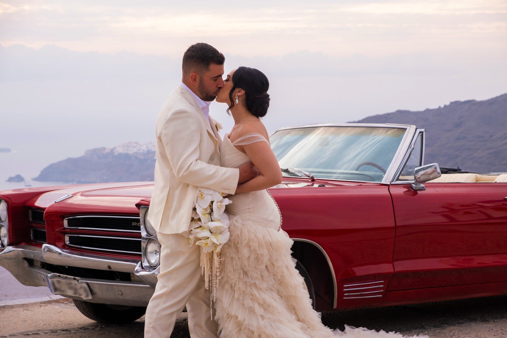 Bride and groom kissing beside the classic 1966 Pontiac - Santorini wedding day photoshoot over the caldera